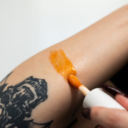 Person applying orange cream to a tattooed arm with a white applicator.