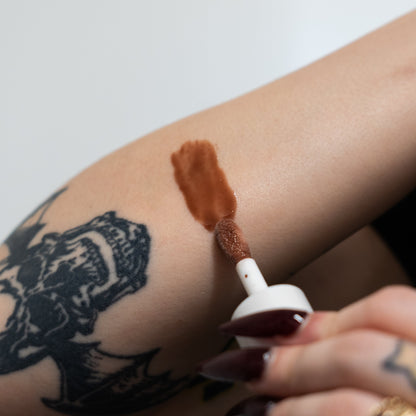 Person applying brown makeup to arm with a brush, close-up on a white background